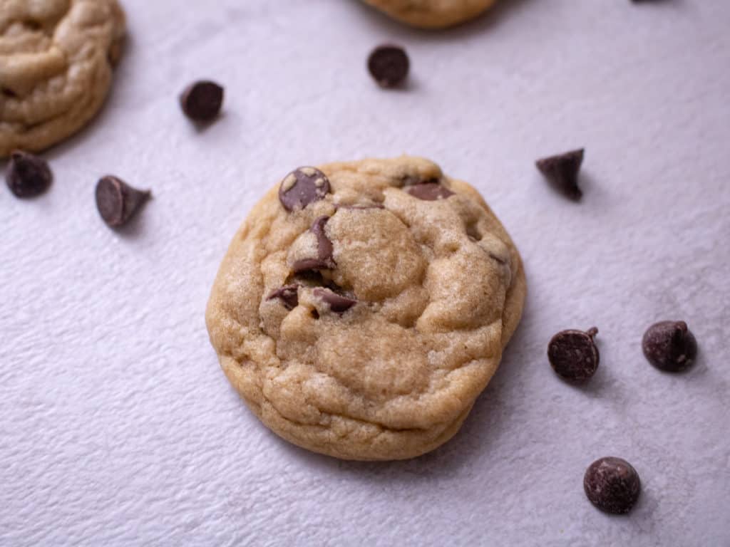 One chocolate chip cookie in the middle of the frame with scattered chocolate chips. 