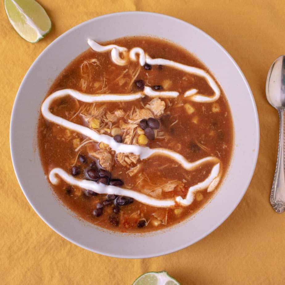 A red, broth based soup with pieces of chicken, corn and black beans visible. Topped with a swirl of sour cream for garnish. On a yellow background. 