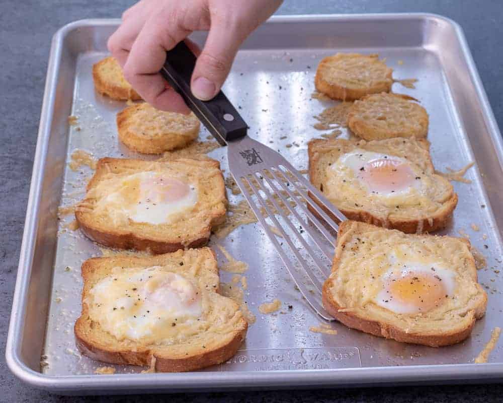 Eggs baked in the middle of toast on a sheet tray. A hand with a spatula is pick up one egg in a hole. There are four total. 