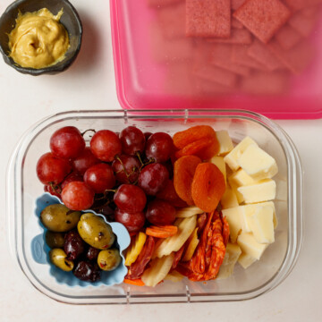 Crackers in a pink reusable bag, mustard in a small brown jar on a white/red background. In a clear glass container is cheese, olives, grapes, dried apricots, deli meat and carrot slices.