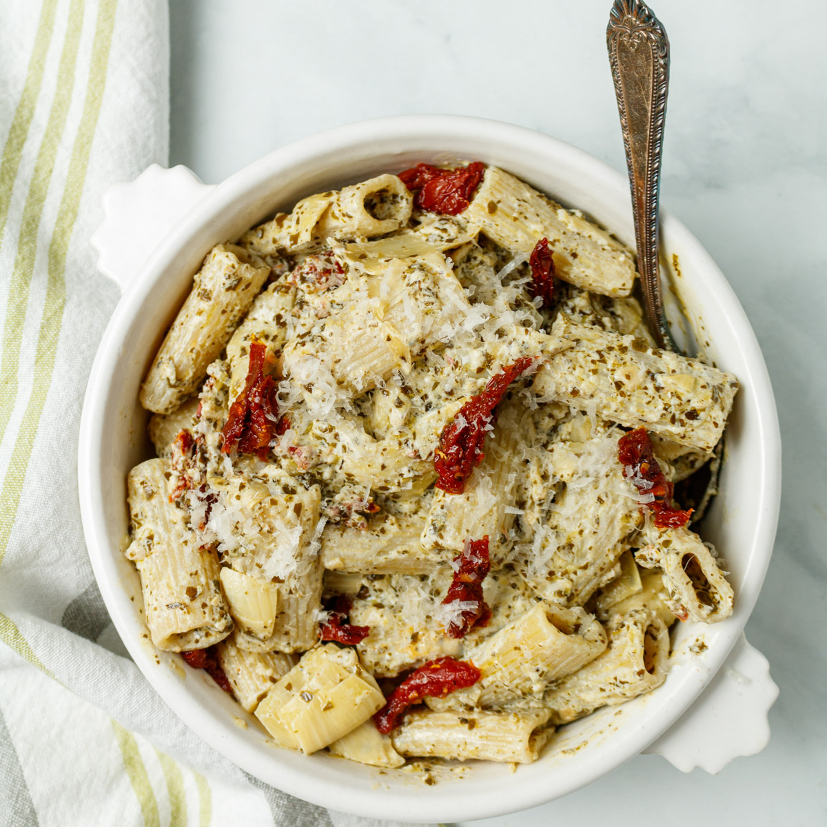 Overhead photograph of pesto pasta in a white bowl with a utensil sticking out from the bowl on the right and a green, white and gray napkin underneath the bowl on the left side.