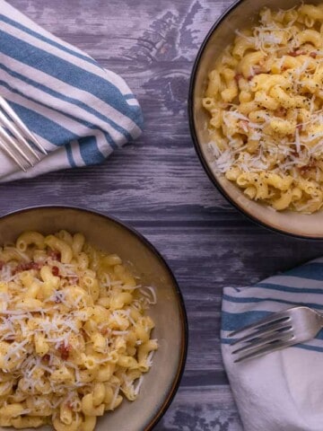 2 bowls of pasta carbonara on a wood background with white and blue napkins.