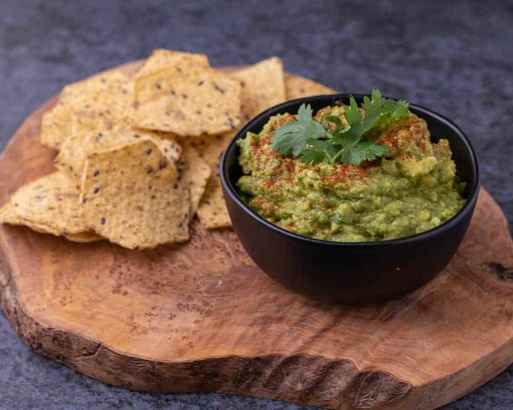 Guacamole in a black bowl topped with paprika and cilantro next to multigrain chips on a wooden platter.