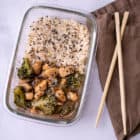 A close up of the meal prep container of chicken and broccoli with rice and oyster sauce. On a brown napkin with wooden chopsticks