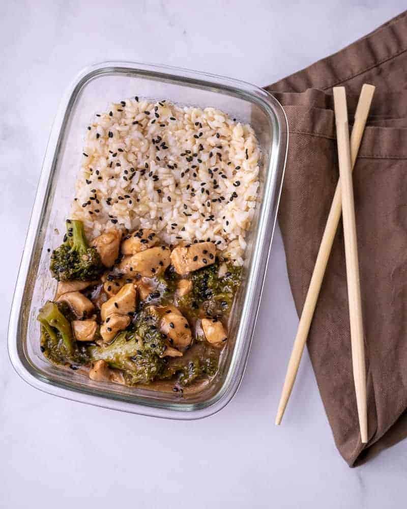 A close up of the meal prep container of chicken and broccoli with rice and oyster sauce. On a brown napkin with wooden chopsticks