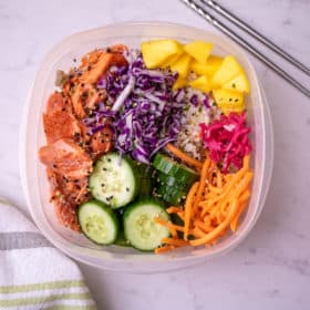 Salmon Poke bowl in a rectangular meal prep container with chop sticks. Featuring cooked salmon, fresh mango, matchstick carrots, pickled ginger, cabbage and cucumbers