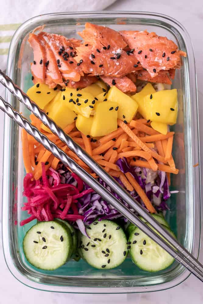 Salmon Poke bowl in a rectangular meal prep container with chop sticks. Featuring cooked salmon, fresh mango, matchstick carrots, pickled ginger, cabbage and cucumbers