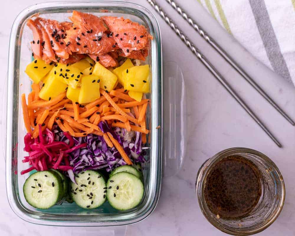 Salmon Poke bowl in a rectangular meal prep container with chop sticks. Featuring cooked salmon, fresh mango, matchstick carrots, pickled ginger, cabbage and cucumbers and sauce on the side.