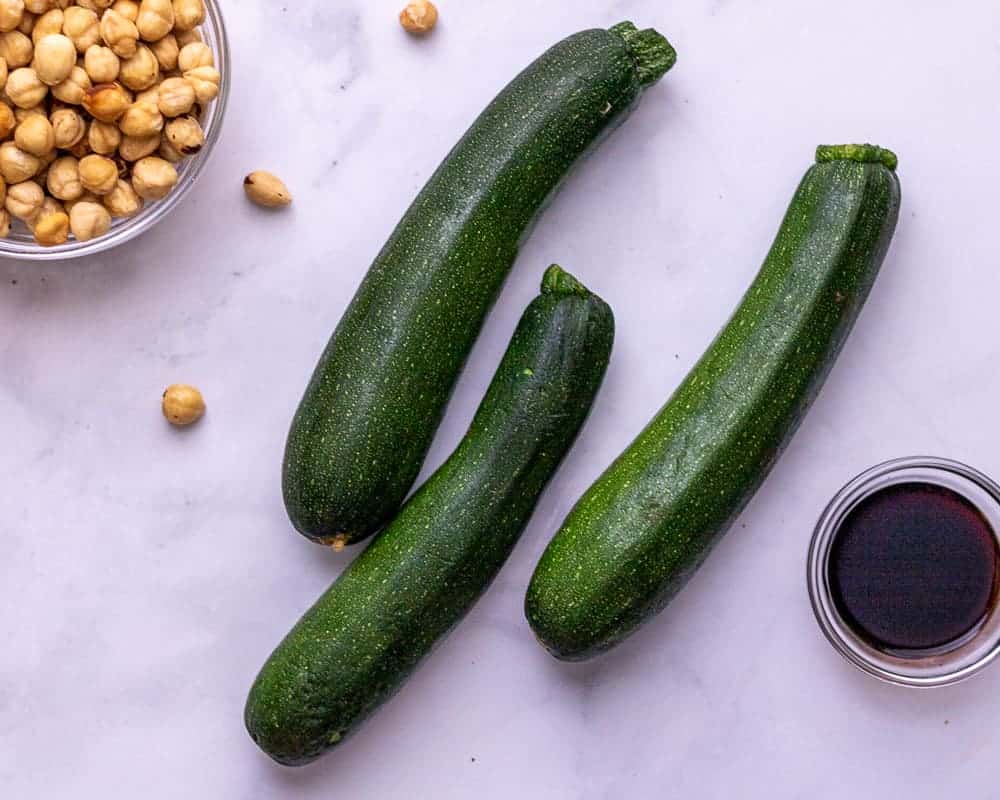 Raw zucchini in the center. A bowl of hazelnuts in the corner and a bowl of balsamic vinegar in the other corner.