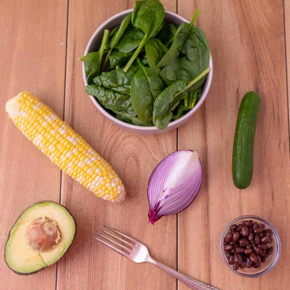 On a wooden background, a bowl of spinach, ear of corn, half an avocado, quarter of a red onion, cucmeber and flake beans.