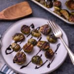 Roasted brussel sprouts on a plate, drizzled with balsamic glaze. A fork is on the plate and a wood spoon is in the frame.