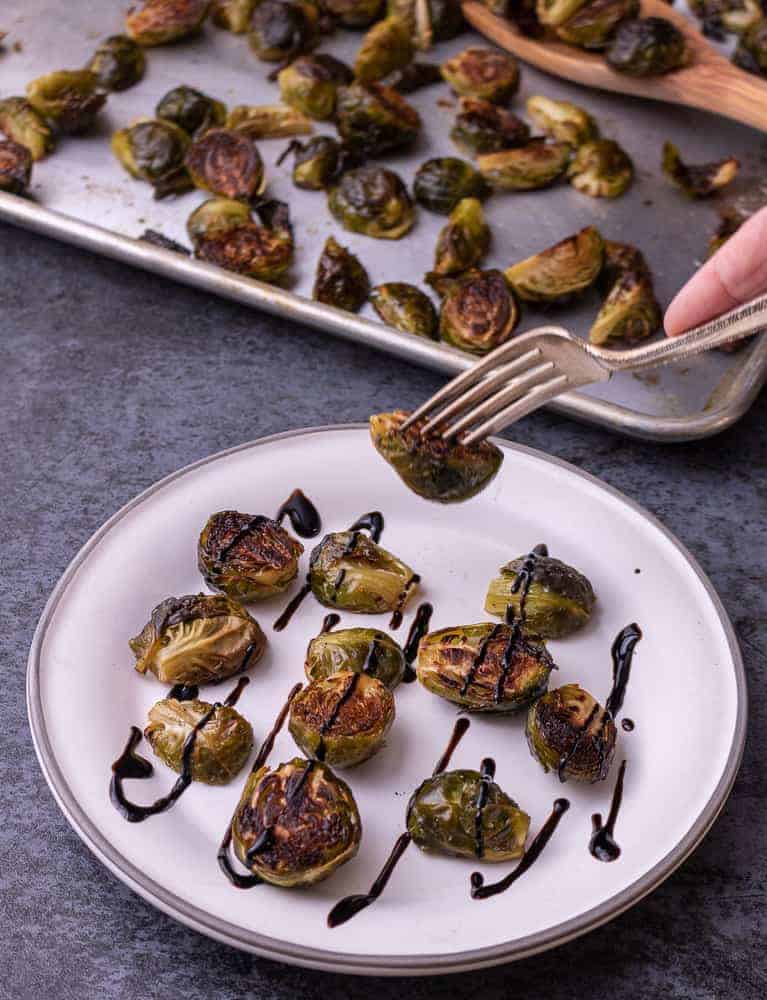 Roasted brussel sprouts drizzled with balsamic reduction. A hand with a fork is picking up a brussel sprout.