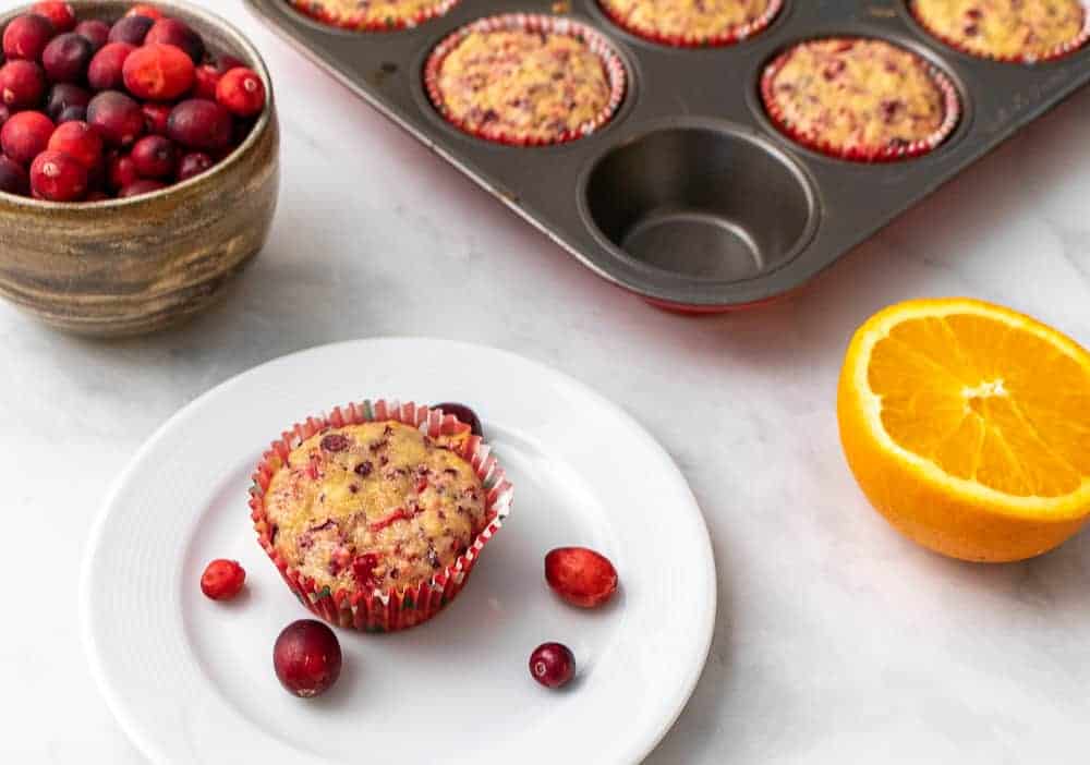One muffin on a white plate in front of a tray of muffins, half an orange and a bowl of fresh cranberries.