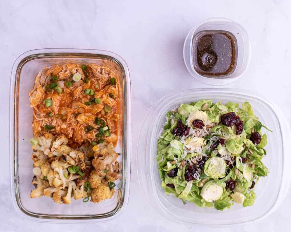 Shredded buffalo chicken in a container with roasted cauliflower next to a food storage container full of brussels sprout meal prep salad. 