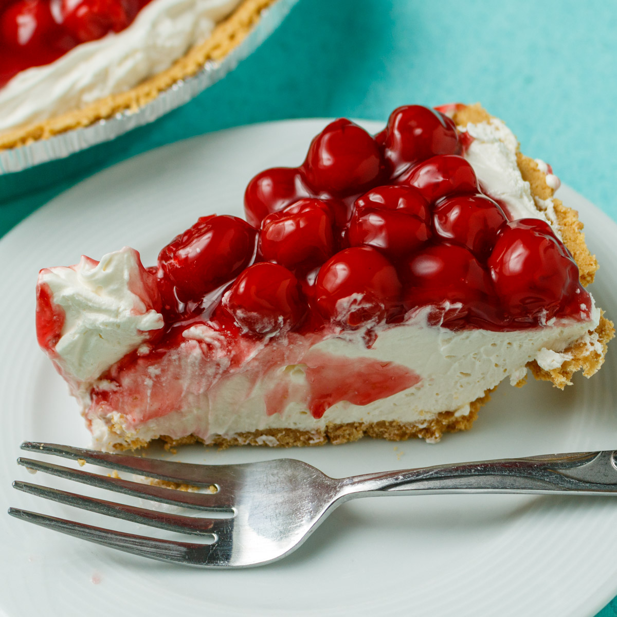 A slice of cherry pie on a white plate with a metal fork in front of it. An edge of the whole pie is seen in the back left corner.