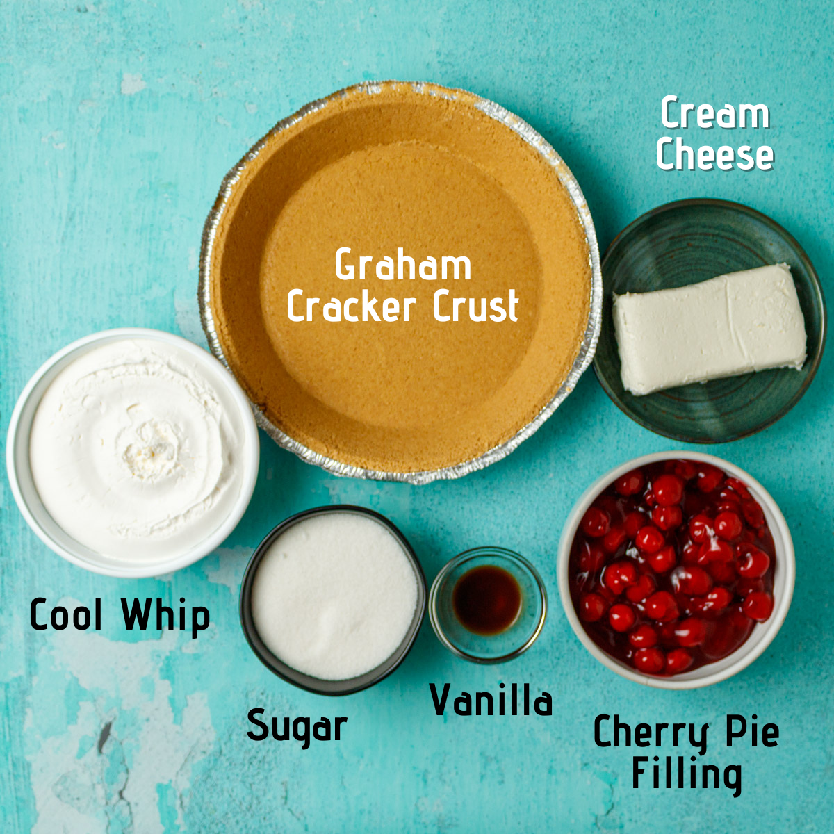 Raw ingredients laid out and labeled on a blue background. Graham cracker crust, cream cheese, cherry pie filling, vanilla, sugar and cool whip.