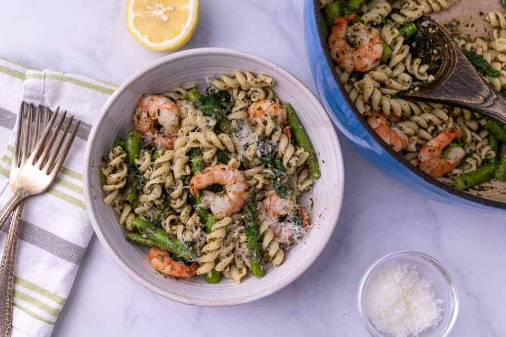 A white bowl of lemon pesto pasta with shrimp next to 2 forks and a dutch oven with parm and lemon in the background