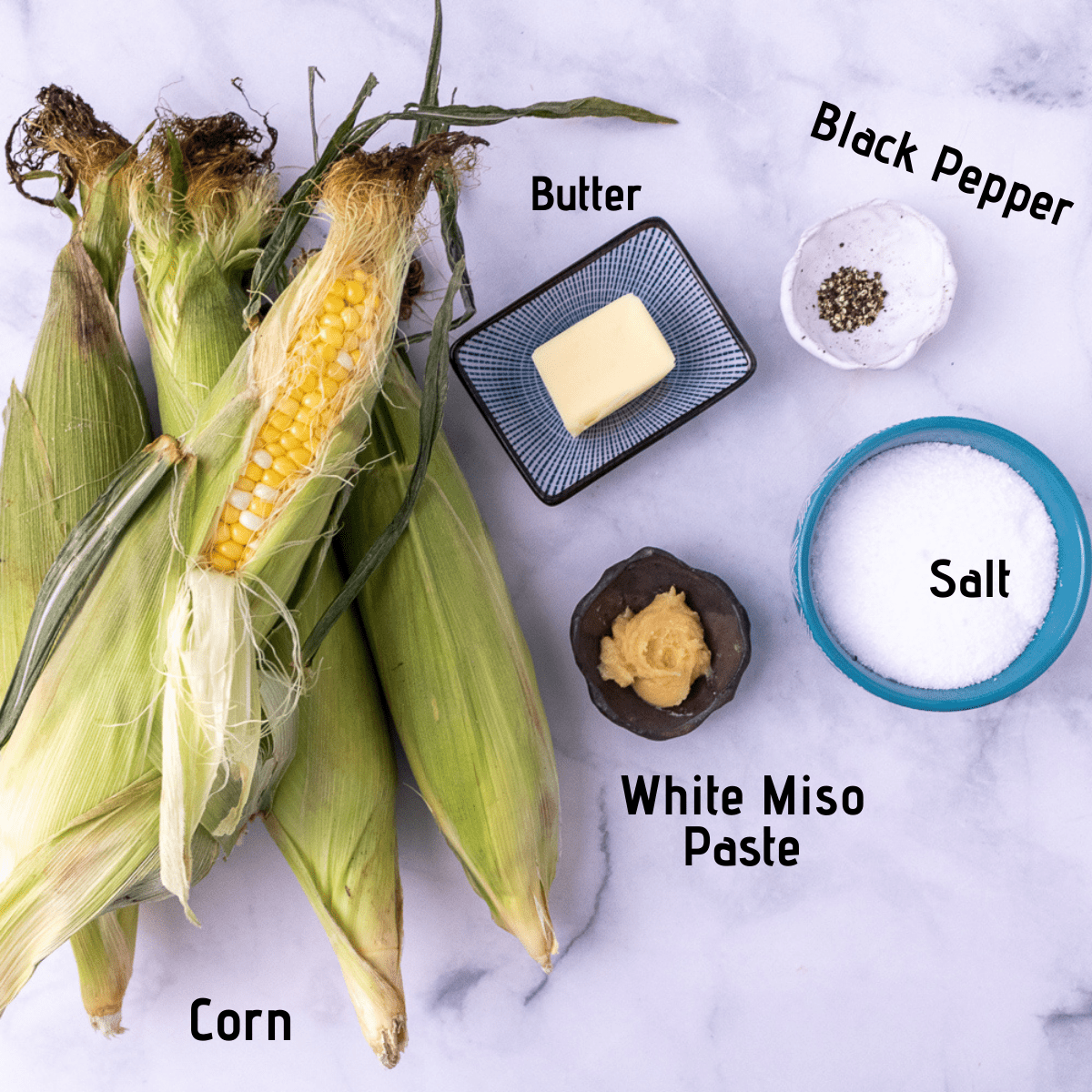 Raw ingredients laid out and labeled on a white marble background, fresh corn, butter, black pepper, salt and white miso paste
