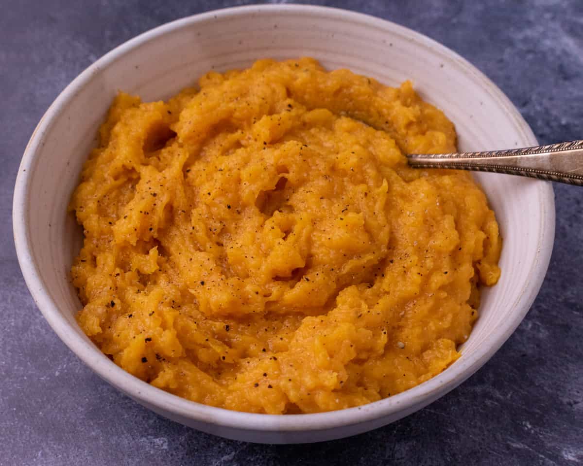 A white bowl of mashed buttter nut squash on a blue background with a spoon