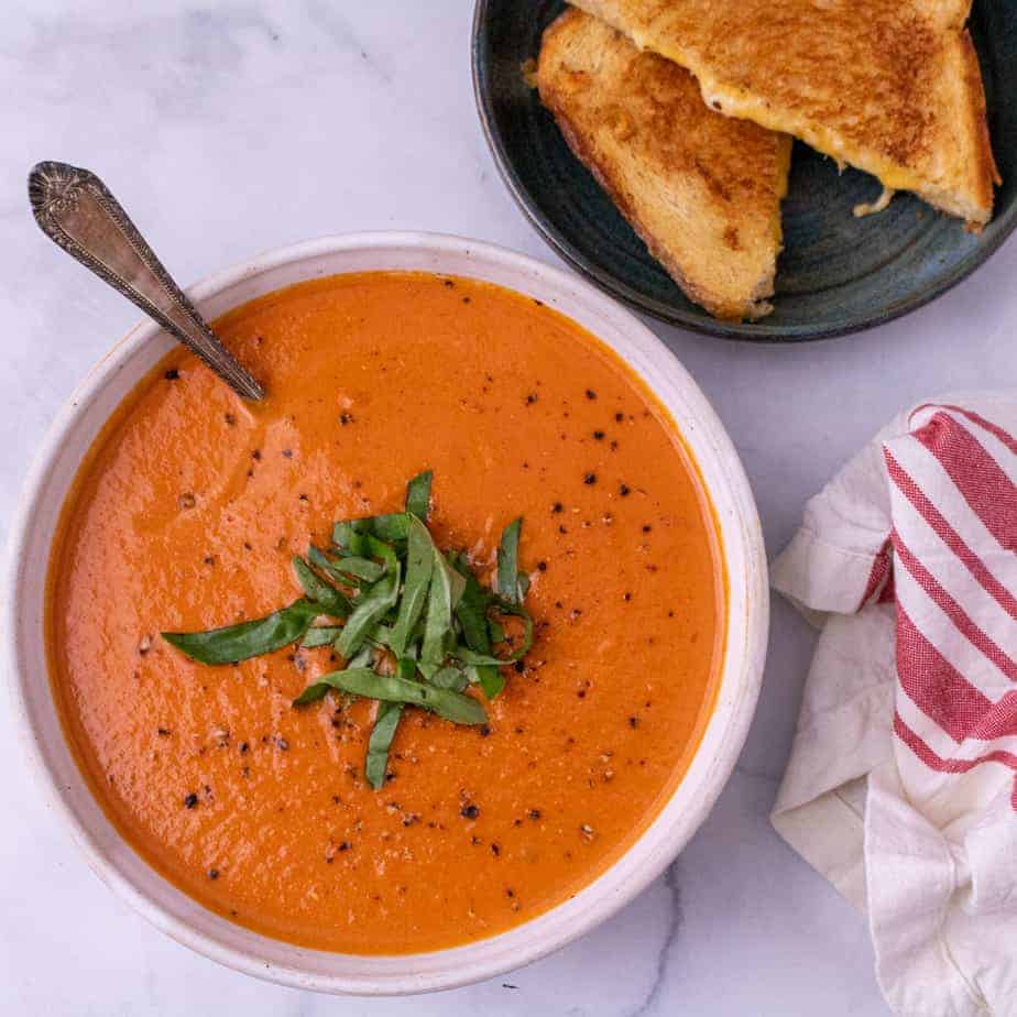 A white bowl of tomato bisque garnished with basil with a spoon. A green plate of grilled cheese is partially visible in the top right corner. A folded red and white napkin is partially visible in the bottom right corner.