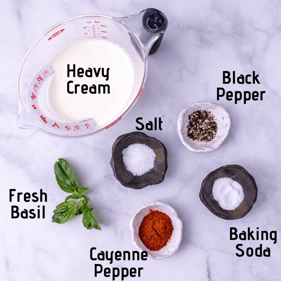 A white background with ingredients laid out with labels; heavy cream, black pepper, salt, fresh basil, baking soda and cayenne pepper.
