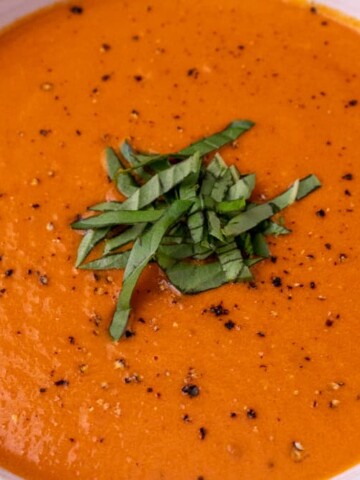 A close up of tomato bisque in a white bowl with black pepper and chopped basil as a garnish