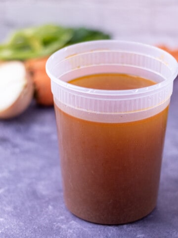 Chicken stock in a plastic container surrounded by carrots, celery, onions and parsley and thyme.