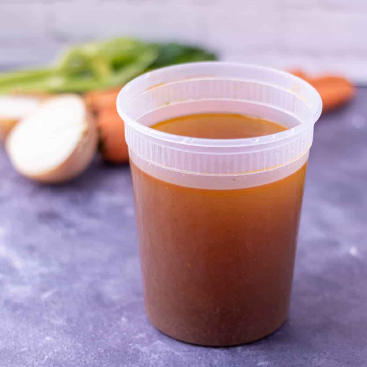 CHicken stock in a plastic container surrounded by carrots, celery, onions and parsley and thyme.