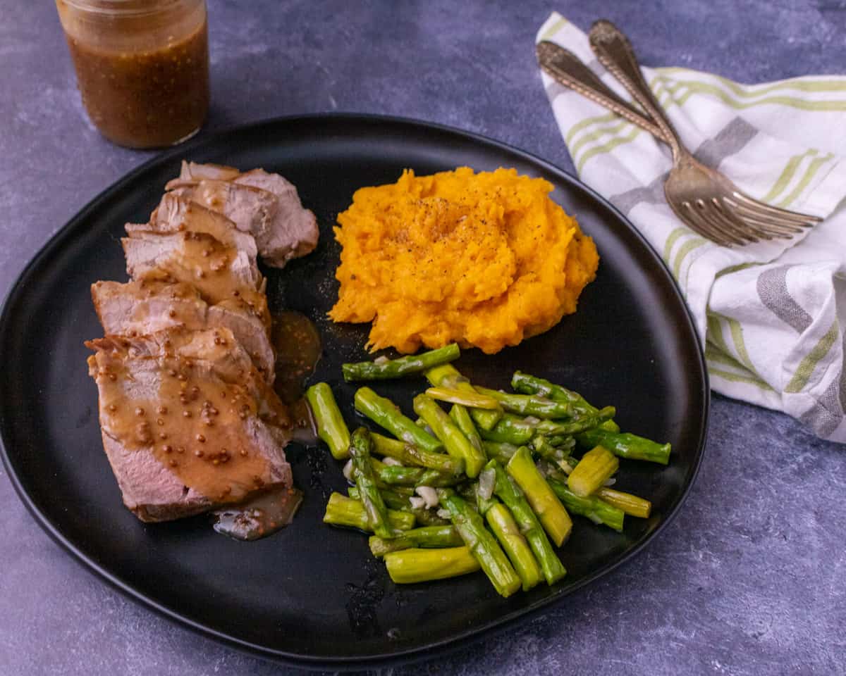 A black plate with sliced pork tenderloin and butternut squash and asparagus. A fork and sauce in a jar in the background.