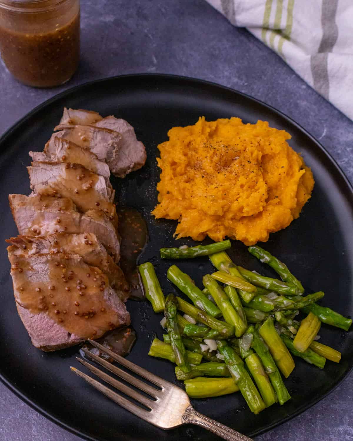 Sliced pork tenderloin with sauce on a black plate with mashed butternut squash, asparagus and a fork.