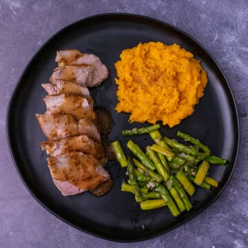 A black plate with sliced pork tenderloin and butternut squash and asparagus.