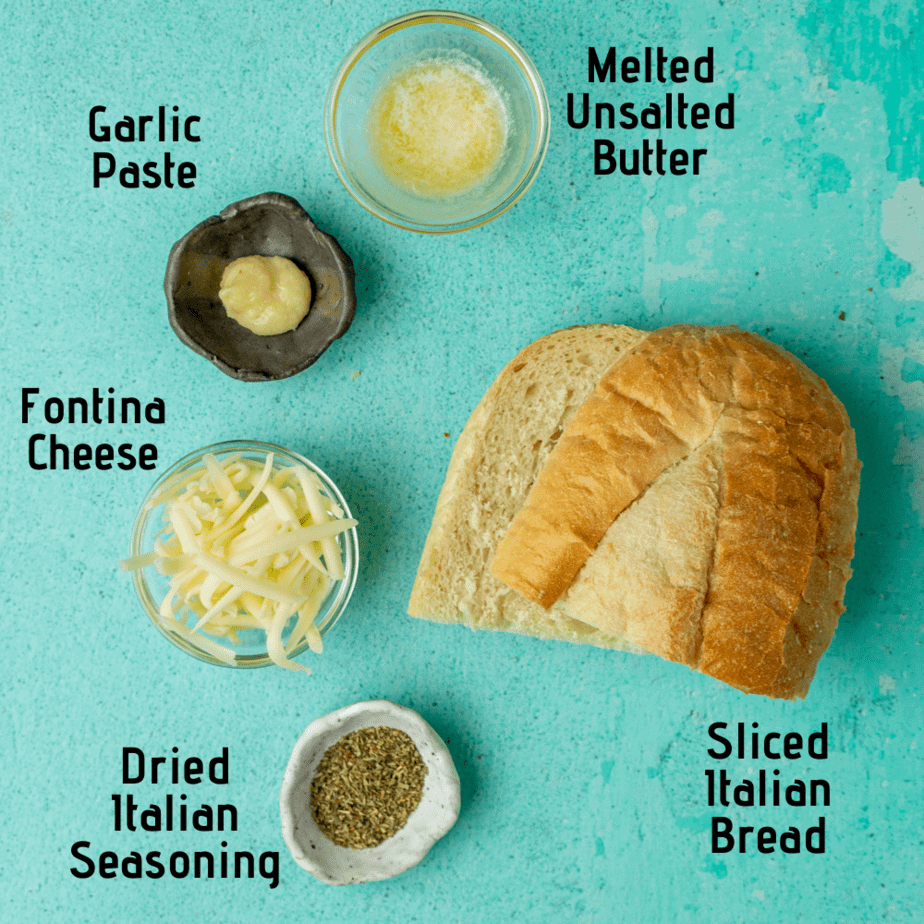 All the ingredients laid out on a blue background and labeled. Garlic paste, melted unsalted butter, sliced italian bread, fontina cheese and dried italian seasoning.