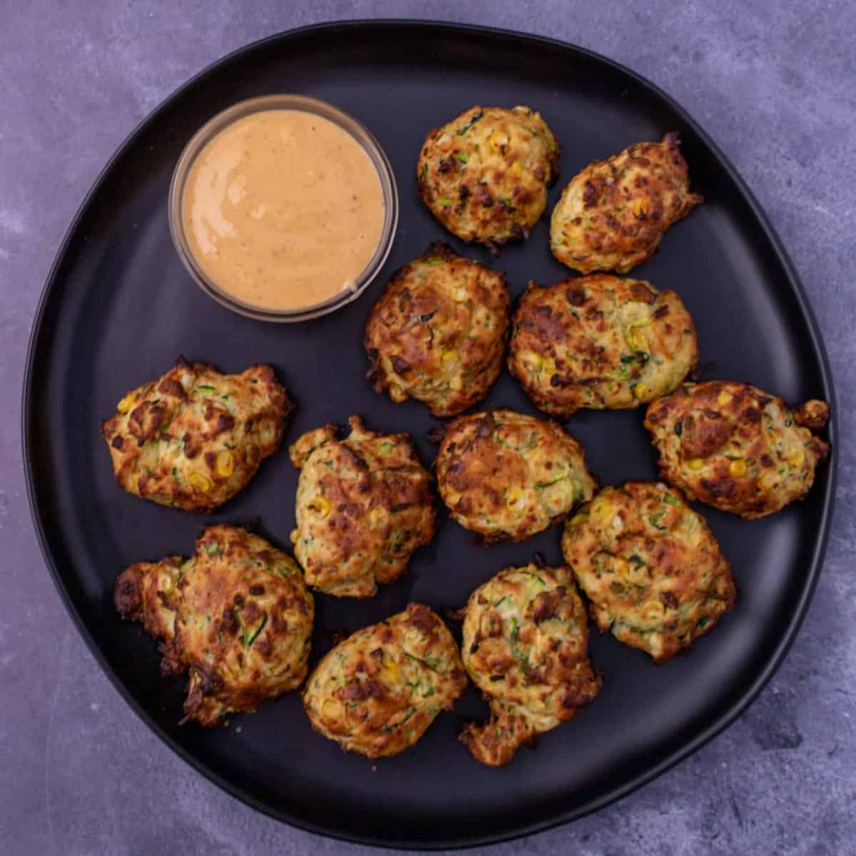 Zucchini fritters on a black plate with sriracha mayo to dip.