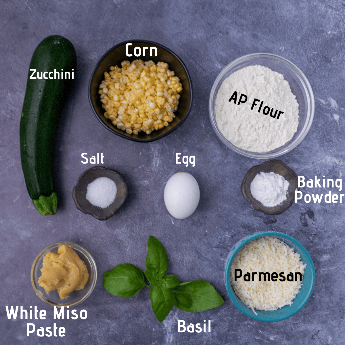 Raw ingredients laid out and labeled, zucchini, corn, AP flour, kosher salt, egg, baking powder, white miso, basil and parmesan