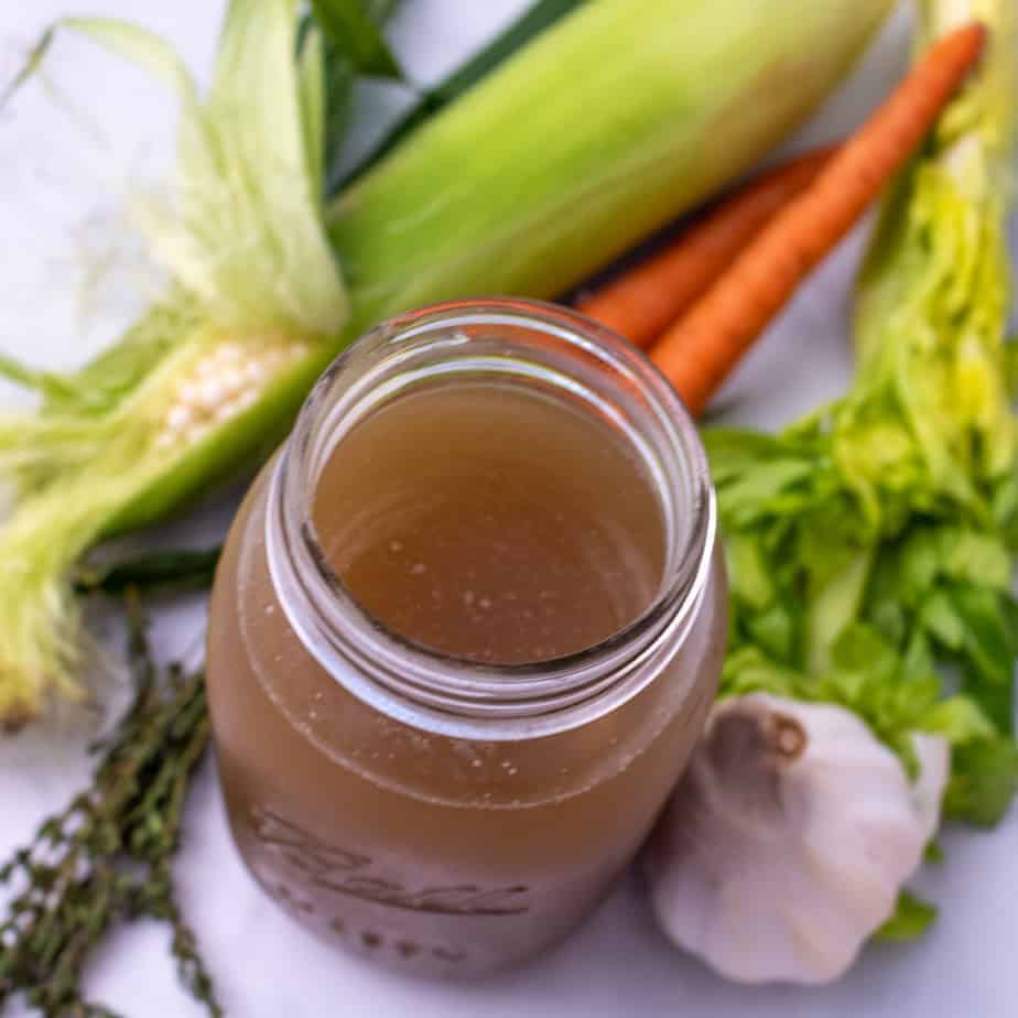 Corn broth in a large glass mason jar surrounded by fresh corn, carrots, celery,thyme and garlic