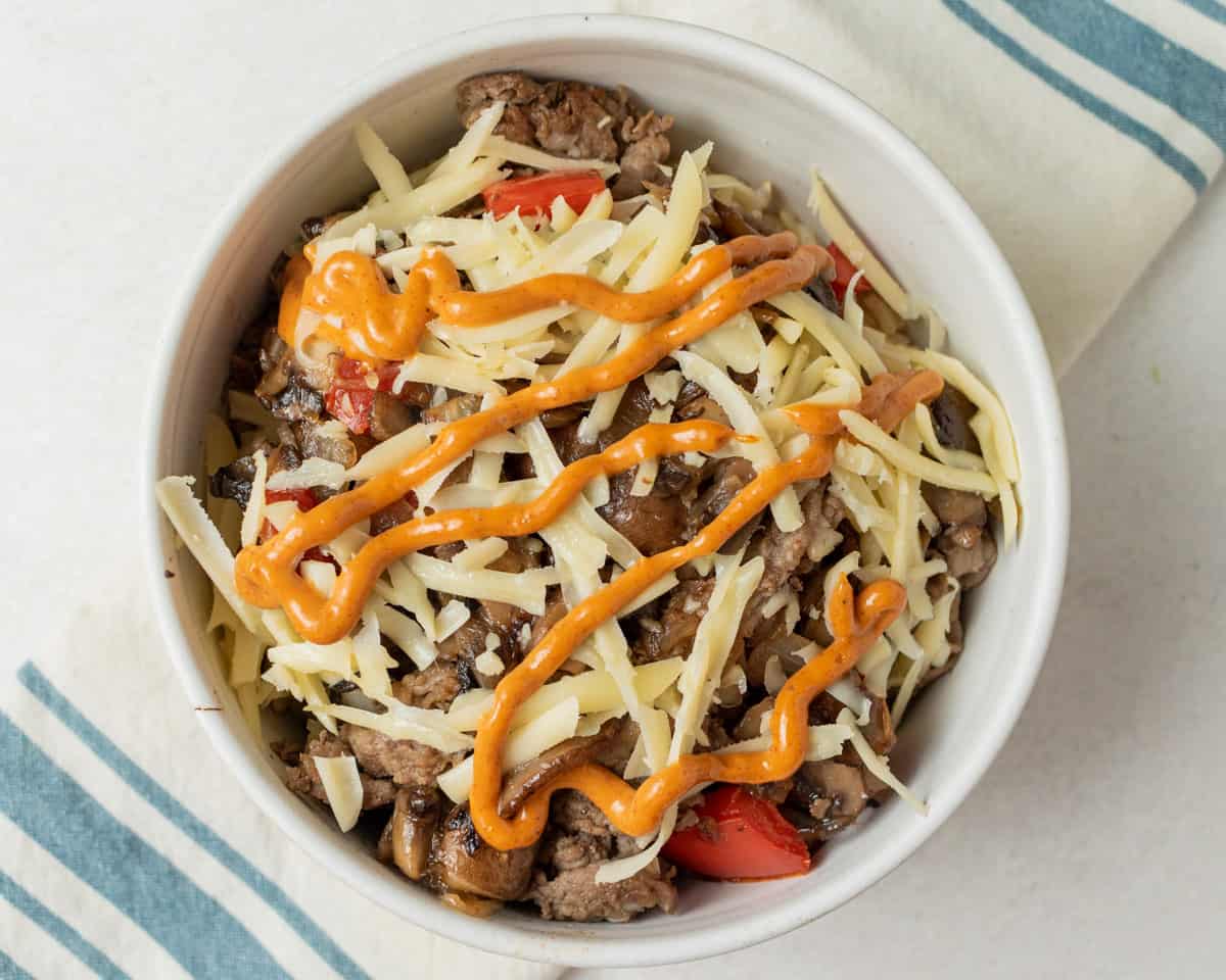 Assembled steak and rice bowl with chipotle mayo drizzled on top. In a white bowl on a white background with a blue striped napkin