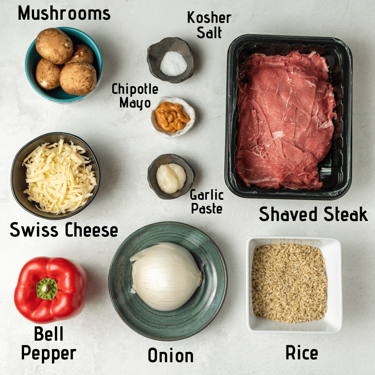 Raw ingredients laid out on a white background and labeled. Mushrooms, kosher salt, chipotle mayo, shaved steak, swiss cheese, garlic paste, bell pepper, onion and rice. 
