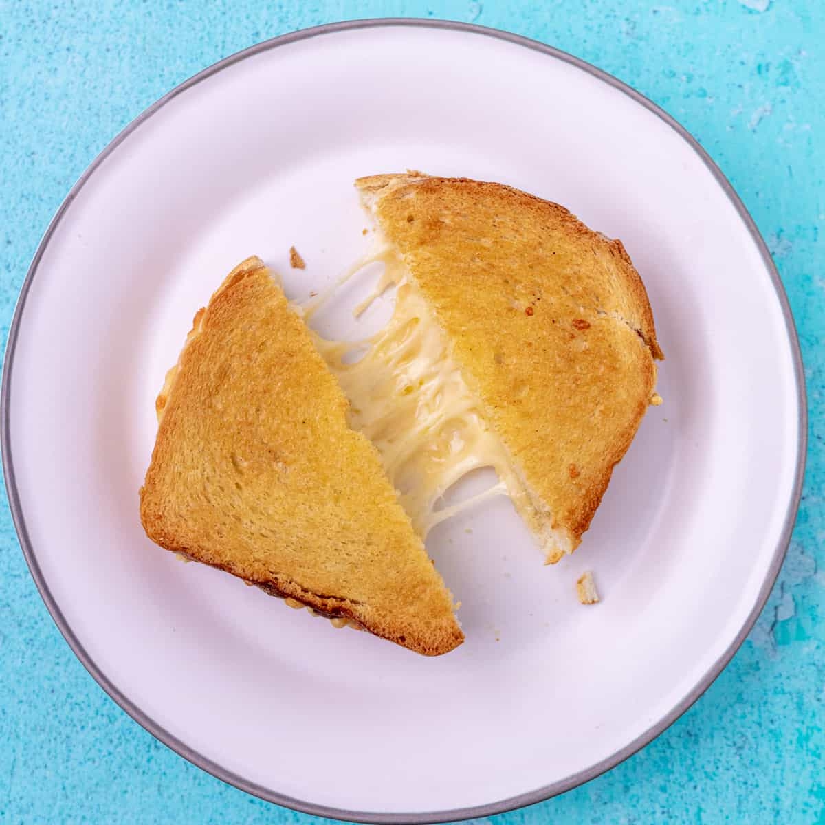 A grilled cheese cut in half with a cheese pull between the two pieces. On a white plate on a blue background.