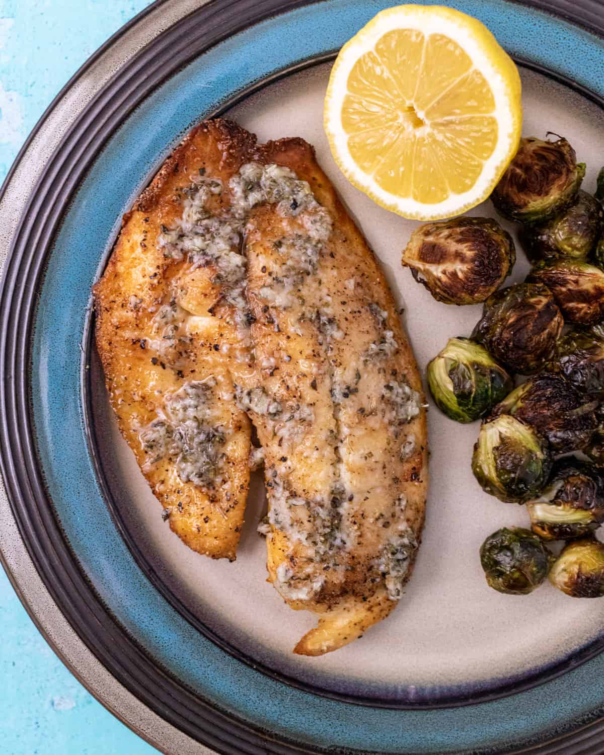 Tilapia on a dinner plate next to roasted brussels sprouts and fresh lemon.