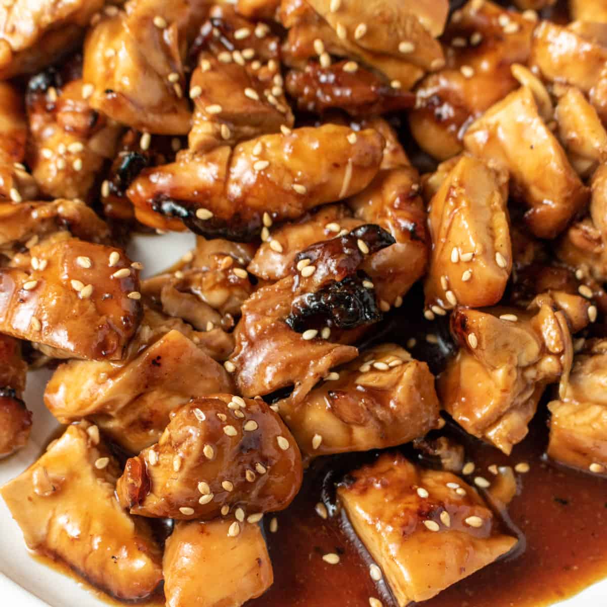 Cut up chicken thighs tossed in teriyaki sauce on a white plate garnished with white sesame seeds.