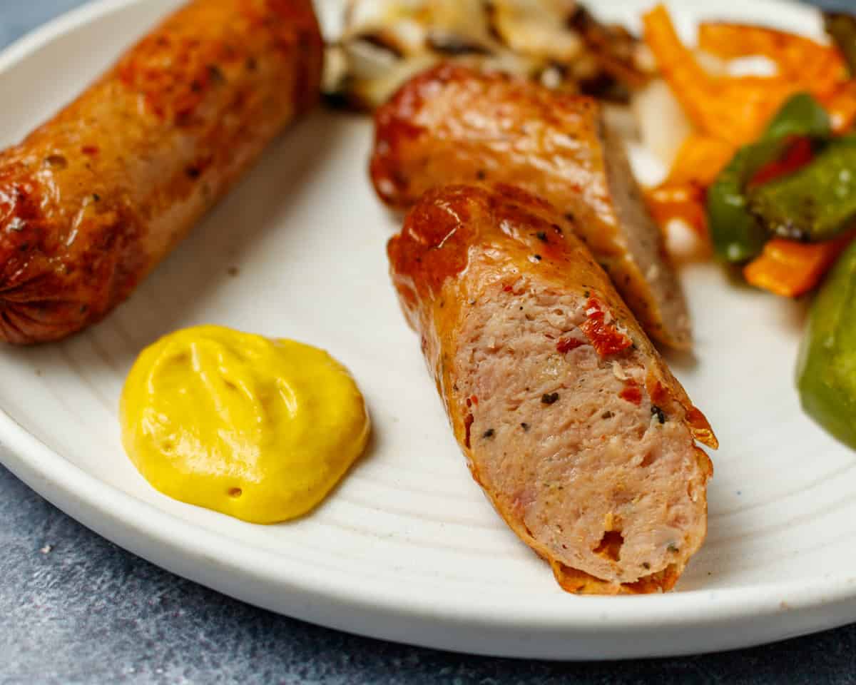 Close up of a cut chicken sausage on a white plate with mustard and vegetables