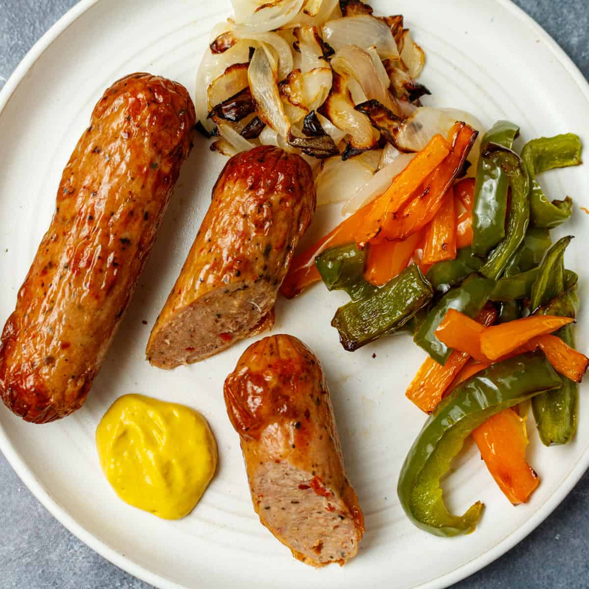 A cut chicken sausage on a white plate with cooked onions, bell peppers and mustard.