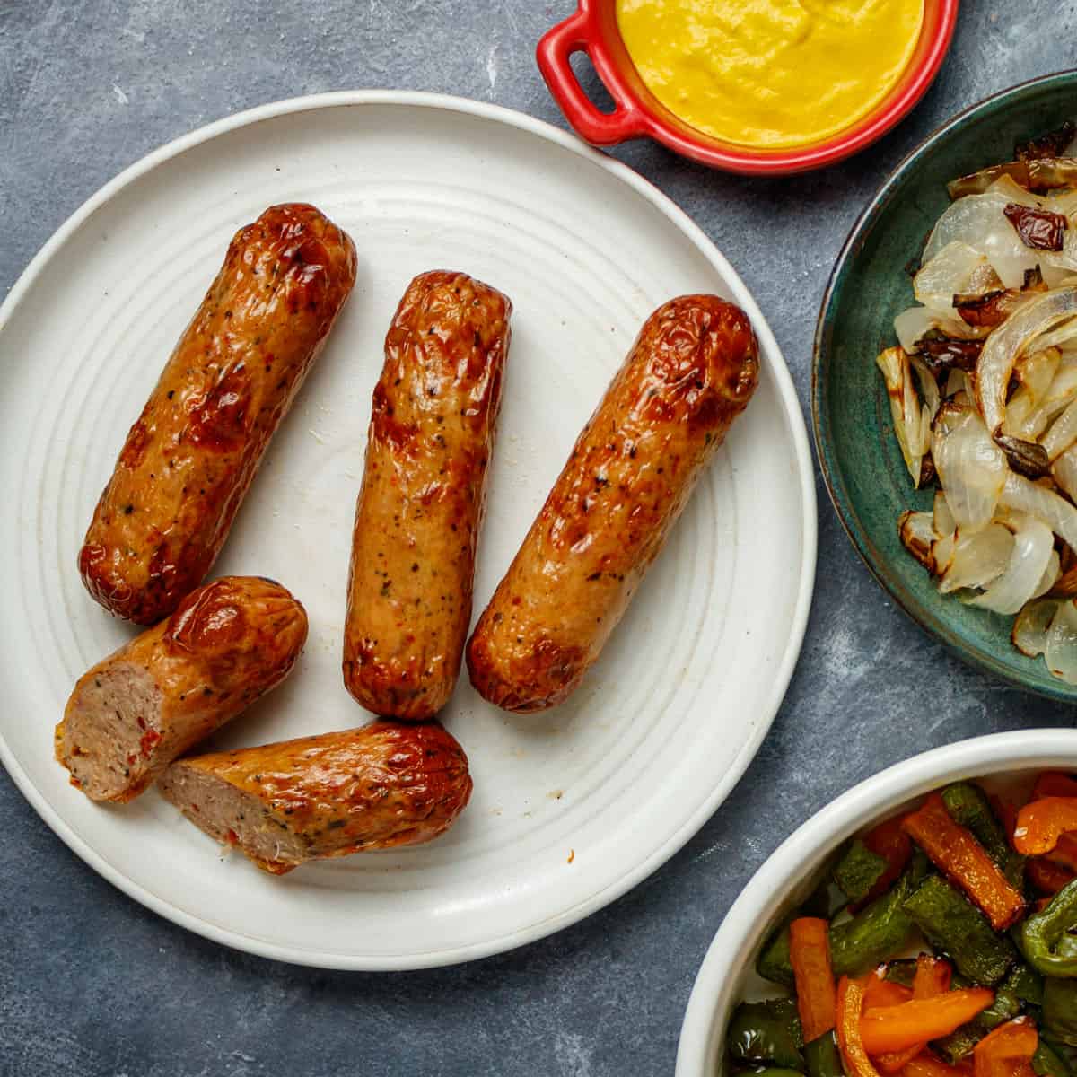 Cooked chicken sausage on a white plate surrounded by mustard, cooked bell peppers and cooked onions.