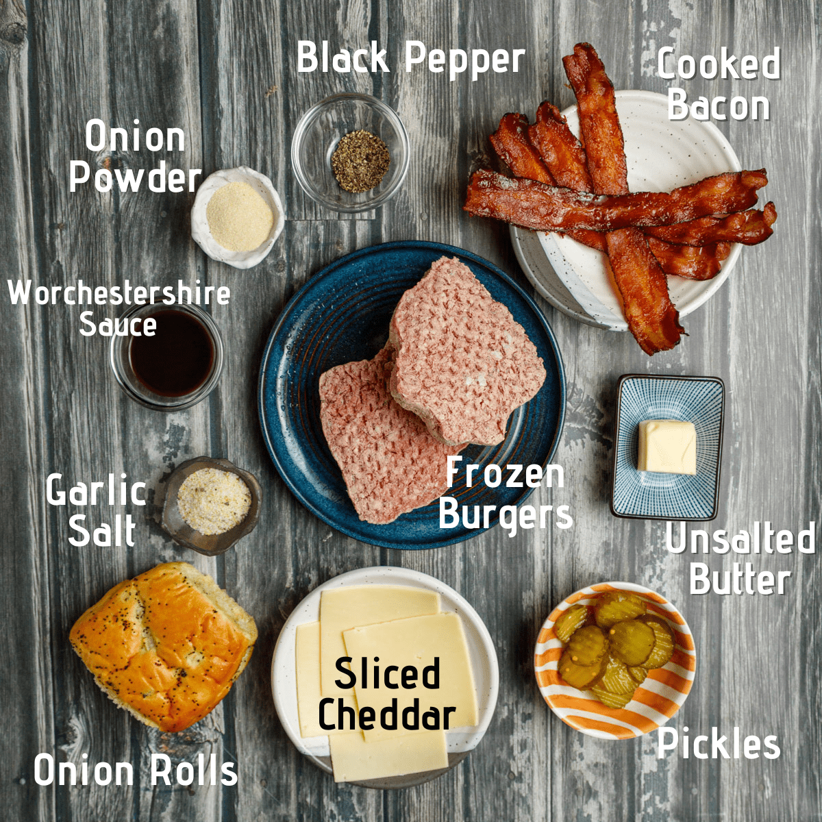 Raw ingredients on a wooden picnic table background. Ingredients are labeled in white and black font: onion powder, black pepper, cooked bacon, unsalted butter, pickles, sliced cheddar, onion rolls, garlic salt, worchestershire sauce and frozen burgers.