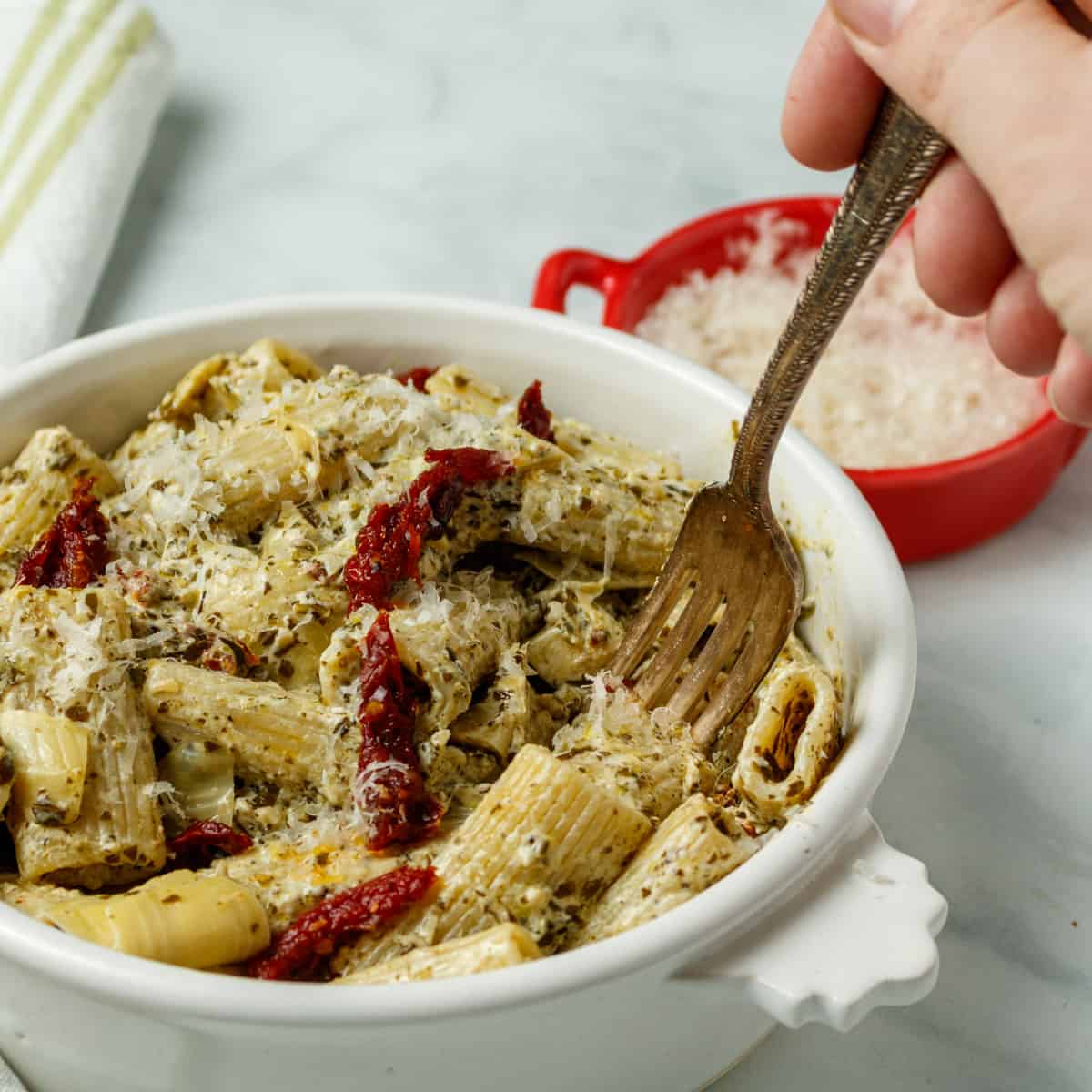 A hand is holding a fork on the right side of the image, stabbing the pasta in a white bowl. In the background is a red bowl with cheese and a white and greennapkin.