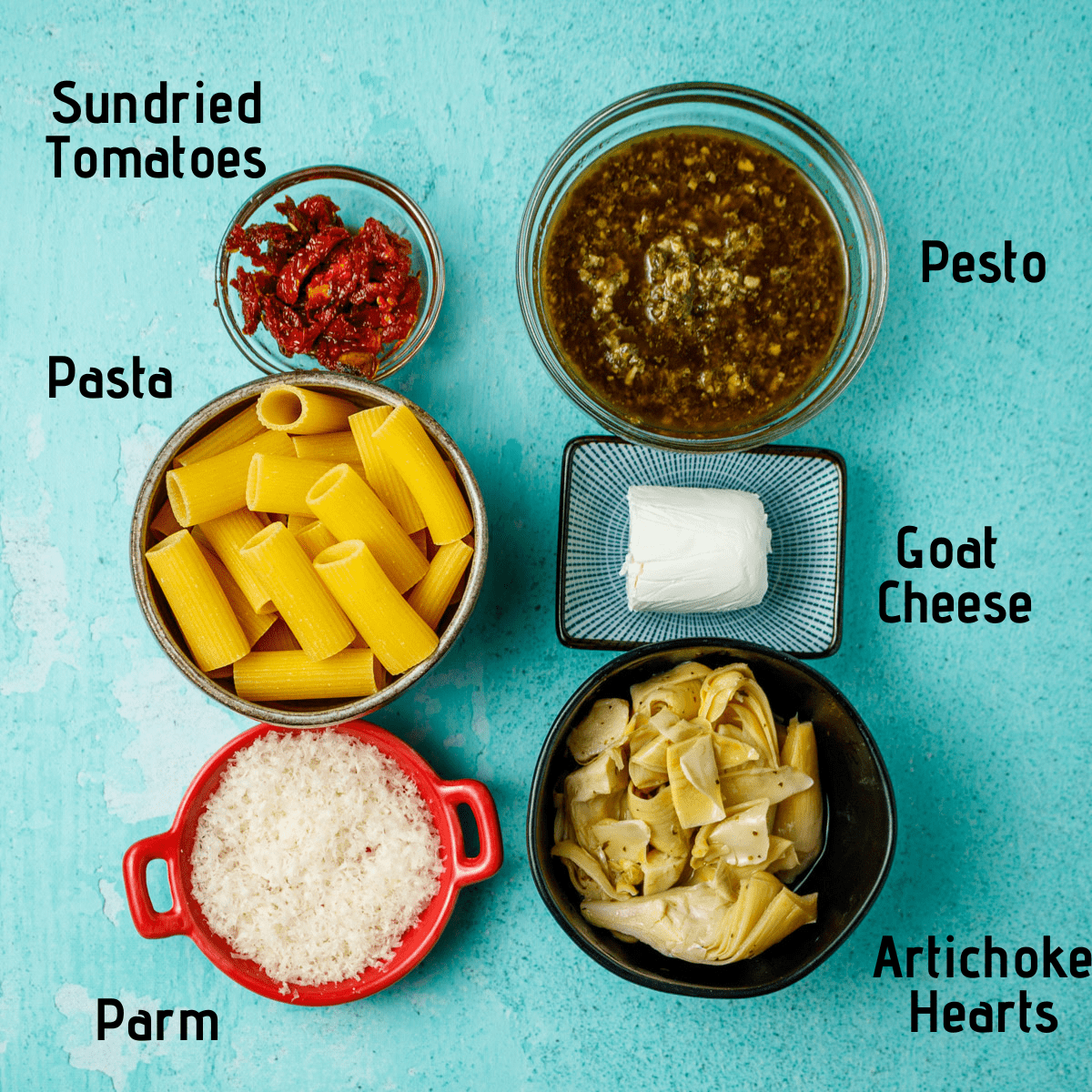 Raw ingredients laid out on a light blue background with black text labels. Sundried tomatoes, pesto, goat cheese, artichoke, parm and pasta.