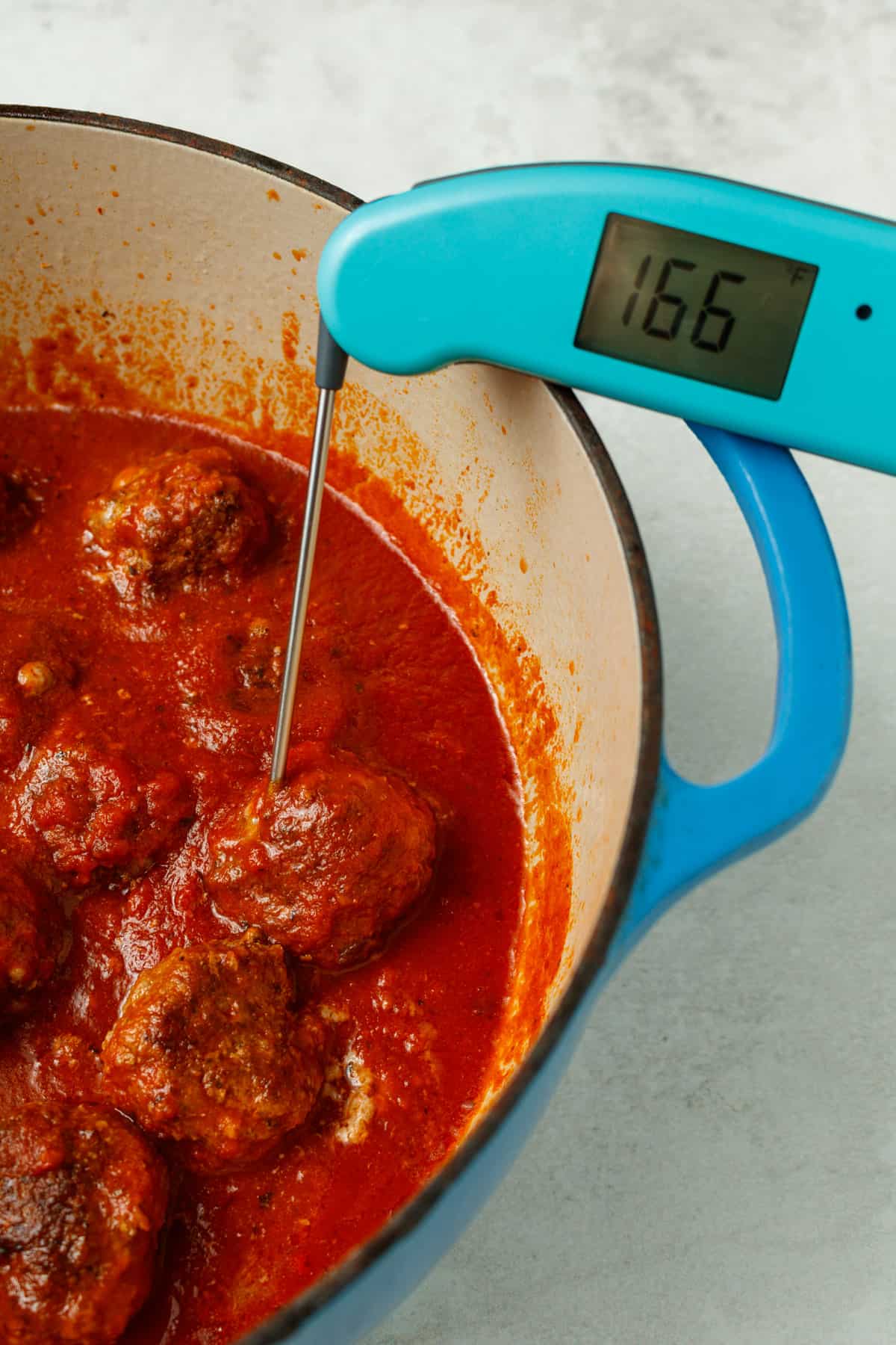 A meat thermometer reading 166F in a meatball. The meatballs are in toamto sauce in a blue dutch oven.
