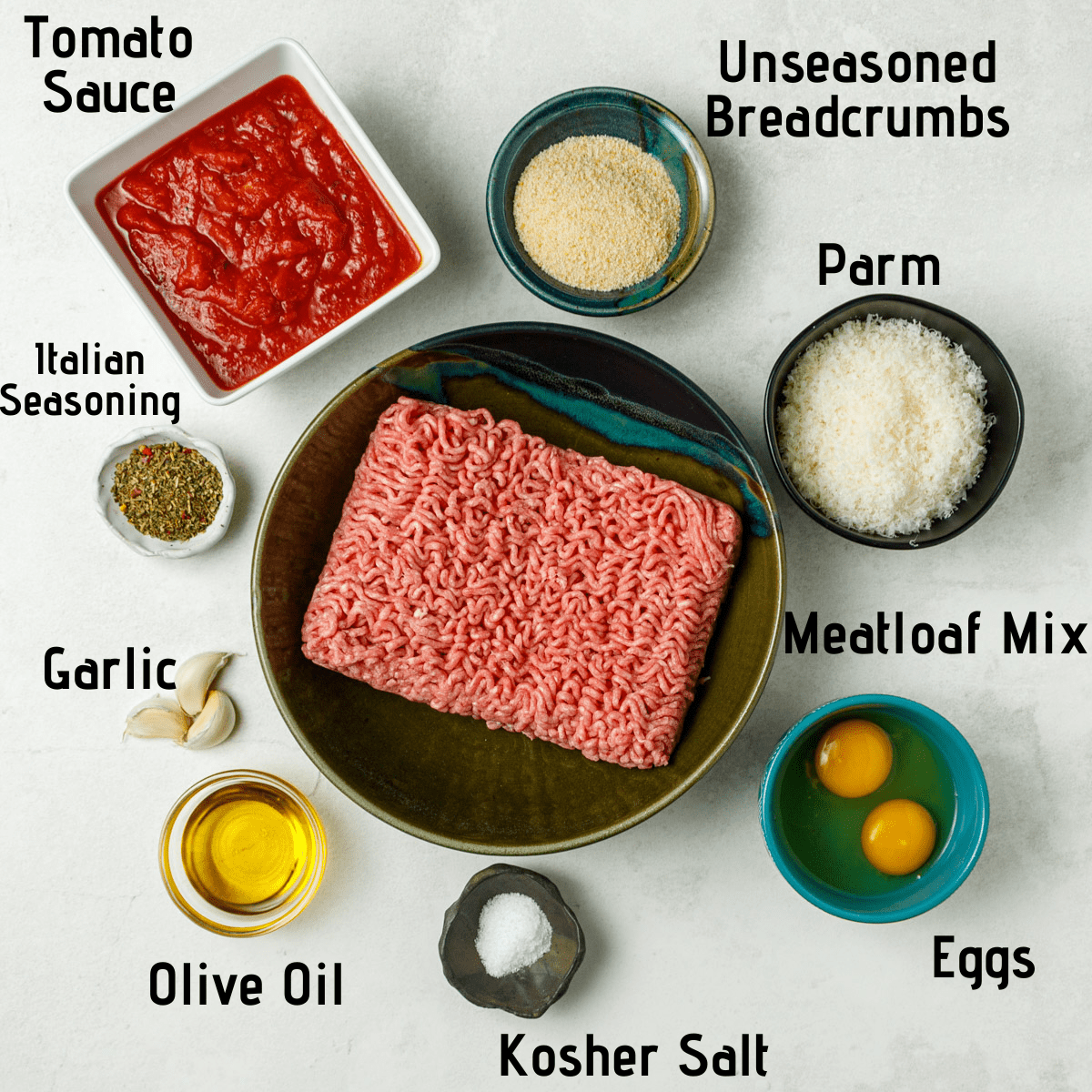 Ingredients laid out on a white background and labeled with black text: Tomato sauce, unseasoned breadcrumbs, parm, meatloaf mix, eggs, kosher salt, olive oil, garlic, italian seasoning