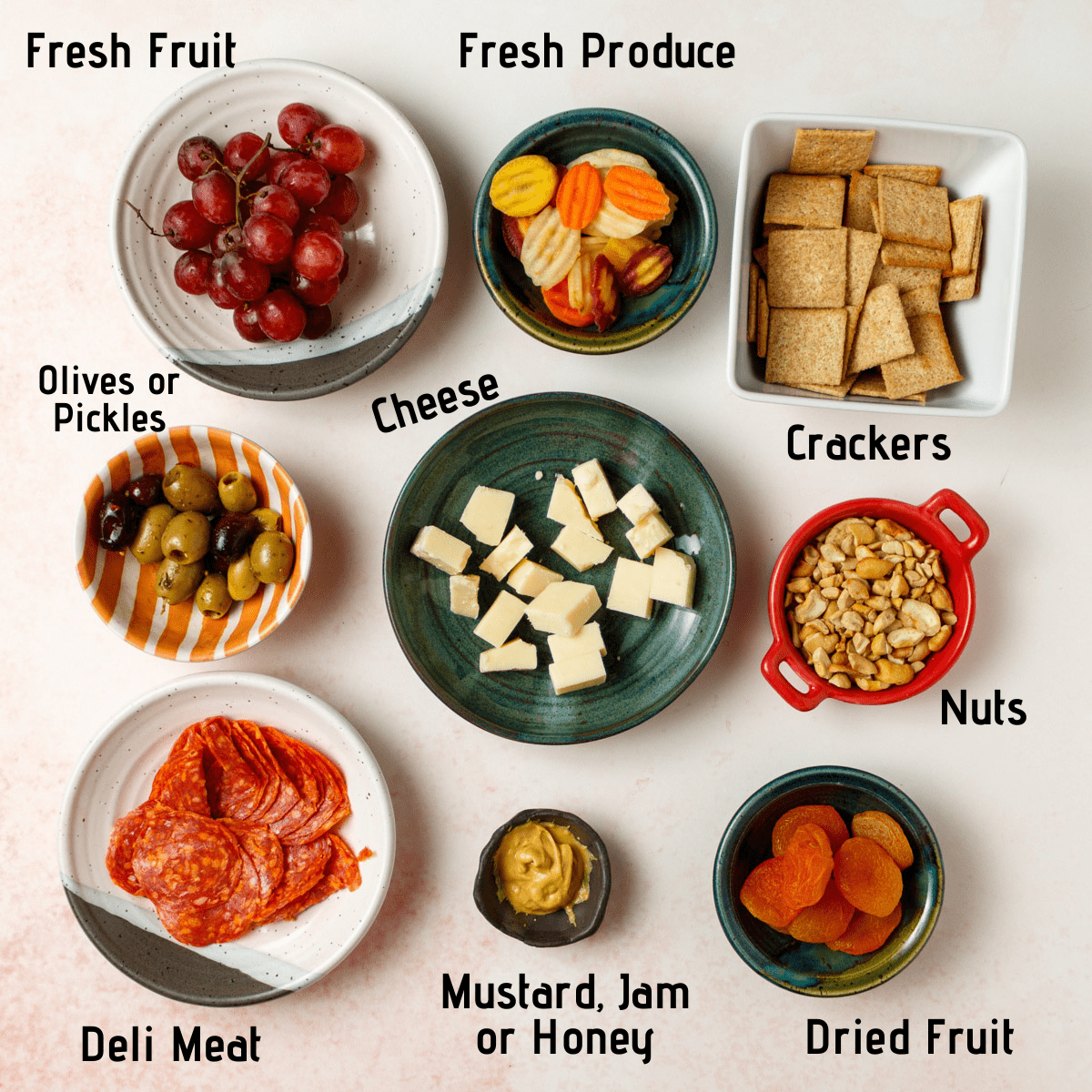 Separate ingredients labeled on a white and red speckled background. Grapes, carrots, crackers, olives, cheese, cashews, deli meat, mustard and dried apricots.